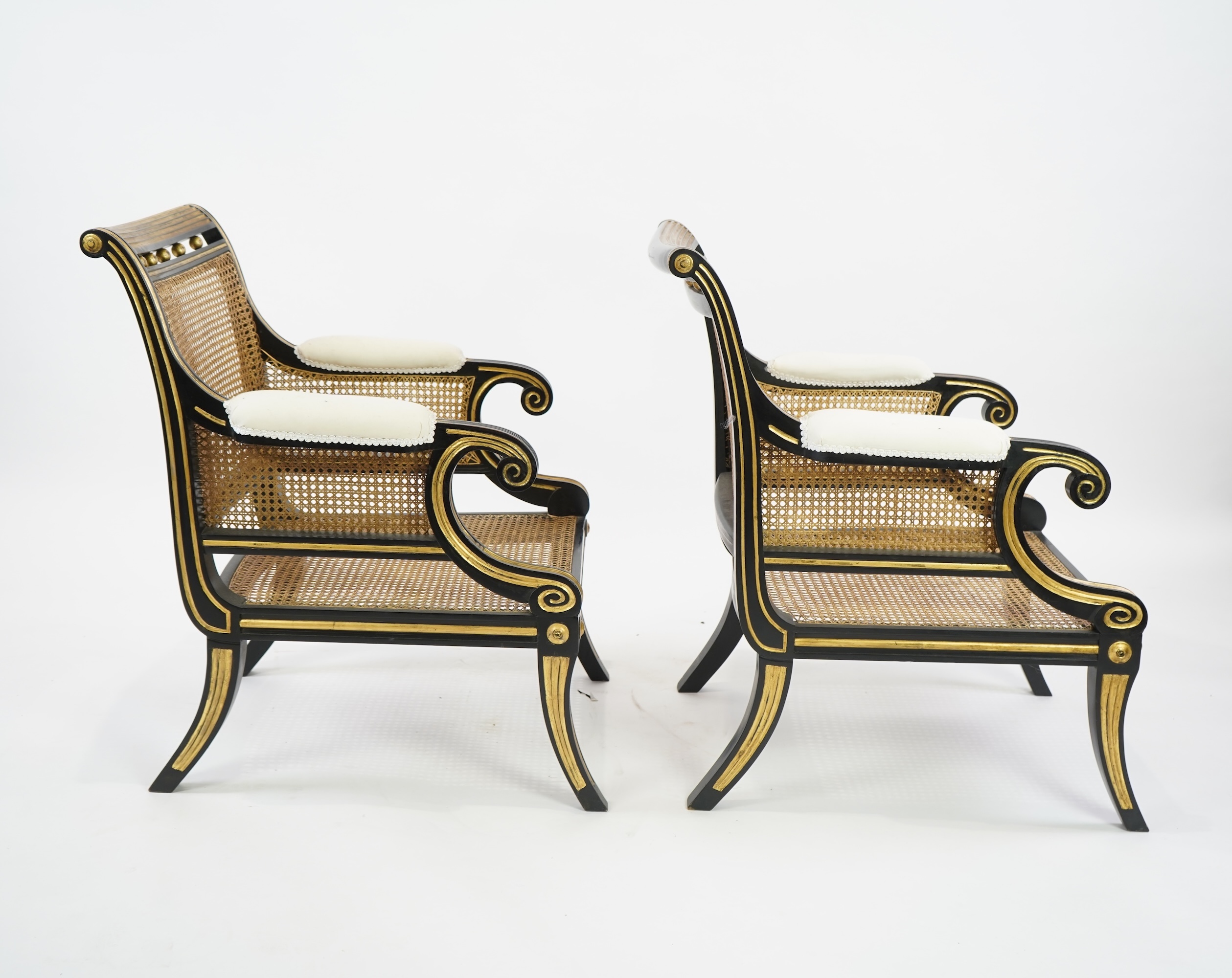 A pair of Regency style ebonised, parcel gilt caned bergere elbow chairs